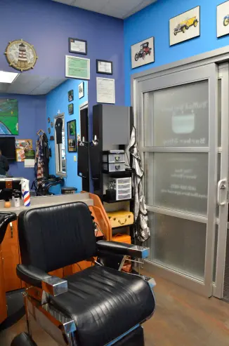 A corner of the barbershop. A barber's chair, a collection of paintings of antique automobiles, and the front door of the barbershop can be seen.